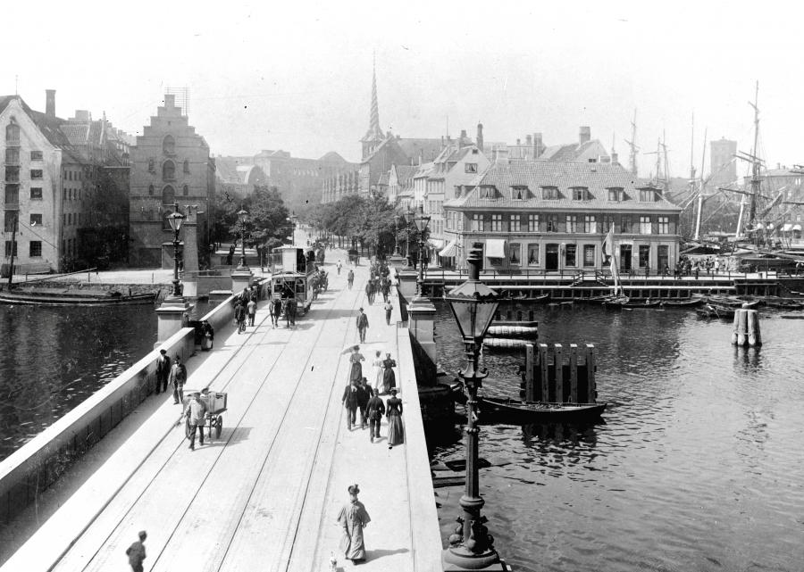 Christianshavnlinien er på vej over Knippelsbro, som i sidste del af 1800-tallet lå i forlængelse af Slotsholmsgade, som ses i baggrunden. Midt i billedet ses spiret på Børsen, og til højre ses tårnet på Nikolaj kirke, inden det fik spir.